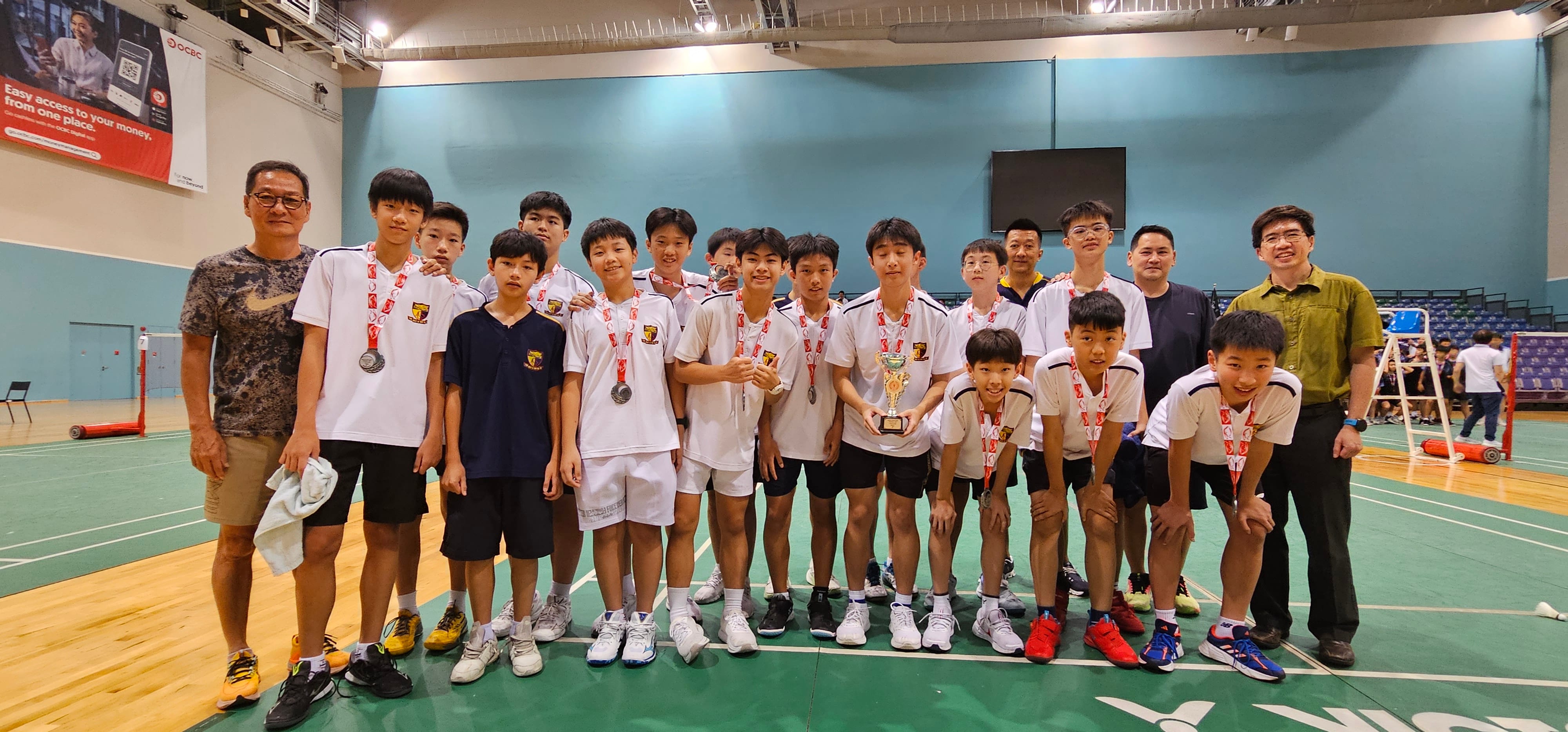 A school badminton team poses with coaches in a sports hall with teal-colored walls, some players wearing medals.